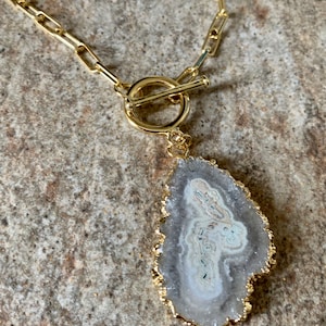 Op de afbeelding: Een gouden ketting met een toggle-sluiting en een grote, grijze agaat hanger met een gouden rand.