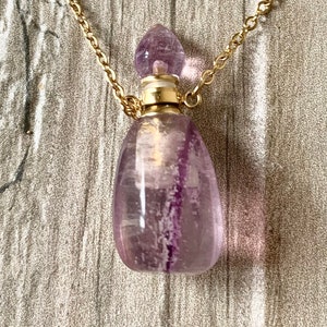 Może przedstawiać: Złoty łańcuszek z wisiorem w kształcie fioletowej butelki z kryształu ametystu. Butelka ma złotą zakrętkę i przezroczyste szklane ciało.