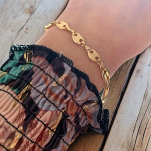 May include: A gold-colored bracelet with a series of oval-shaped links. The bracelet is worn on a wrist, and the background includes a patterned sleeve and a wooden surface.