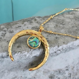 May include: Gold-coloured crescent moon pendant necklace with a hammered texture. The pendant features a round, iridescent abalone shell inlay. The necklace has a delicate chain.
