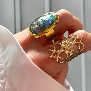 Puede incluir: Dos anillos de oro. Uno presenta una concha de abulón ovalada engastada en una banda de oro. El segundo anillo es un diseño de filigrana dorada. Los anillos se llevan en un dedo, con una camisa blanca en el fondo.