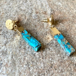 May include: A pair of gold-coloured stud earrings. Each earring features a rectangular turquoise and gold-flecked pendant, and a textured, circular stud. The turquoise pendants have gold-coloured caps and are rectangular in shape.