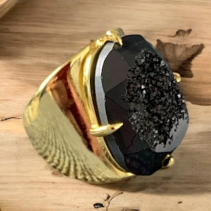 May include: A gold ring featuring a large black agate gemstone. The gemstone has a rough, textured surface and is set in a gold band with a claw setting.