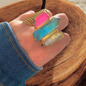 May include: A gold-toned ring featuring three rectangular, colorful stones: pink, blue, and white. The ring has a textured band and a decorative gold setting. The ring is worn on a finger, with a denim sleeve in the background.