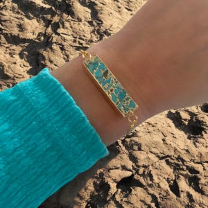 May include: Gold-toned bracelet featuring a rectangular turquoise stone inlay. The bracelet has a delicate chain and a rectangular turquoise stone, set in a gold-colored frame. The turquoise stone has a mosaic pattern. The bracelet is worn on a wrist.