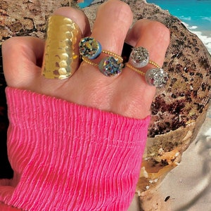 May include: A hand wearing a collection of rings. A large, hammered gold ring adorns the index finger. Two delicate rings feature sparkling, iridescent stones. The rings are set against a backdrop of a pink sleeve and a beach scene.
