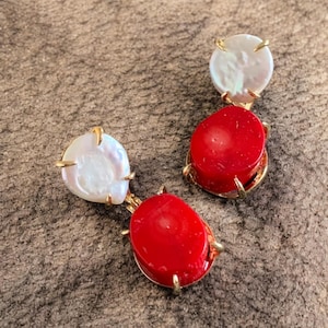 Può includere: Un paio di orecchini dorati con una perla bianca di forma irregolare e una pietra rossa ovale. Gli orecchini hanno una montatura dorata con piccole griffe. Gli orecchini sono esposti su una superficie strutturata di colore neutro.