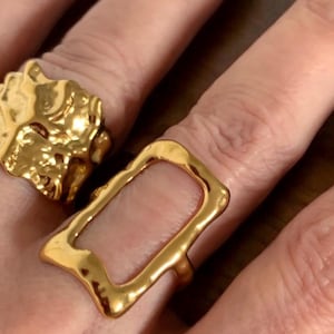May include: Two gold rings on a hand. One ring is a chunky, textured design and the other is a simple, open rectangle.