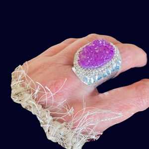 Può includere: Un anello d'argento con una grande pietra preziosa viola scintillante incastonata in una montatura d'argento. L'anello è indossato al dito di una persona.