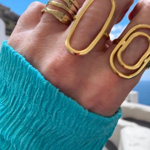 May include: Close-up of a hand adorned with multiple gold rings. The rings feature various designs, including a layered band and elongated oval shapes. The person is wearing a turquoise blue top, creating a contrast with the gold jewelry.