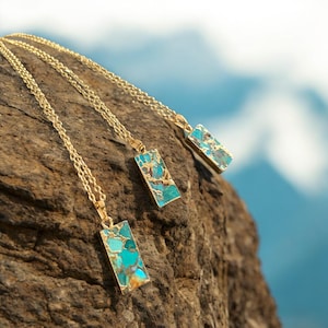 May include: Three gold-colored necklaces with rectangular pendants. The pendants feature a turquoise and brown stone set in a gold frame. The necklaces are displayed on a textured, brown surface with a blurred blue background.