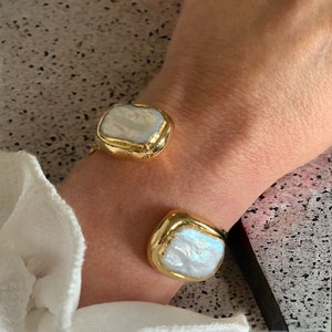 May include: Gold-tone cuff bracelet featuring two large, irregular-shaped, iridescent white pearls. The pearls are set in a textured gold metal. The bracelet is open, allowing it to be adjusted to fit the wrist.