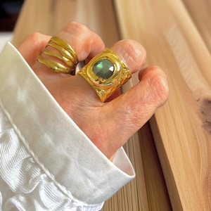 May include: Two gold rings are shown on a hand. One ring has a layered design, and the other is a square shape with a green gemstone. The rings are set against a light background.