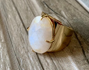 May include: A gold ring featuring a large, oval-shaped moonstone set in a claw setting. The ring has a thick, wide band.