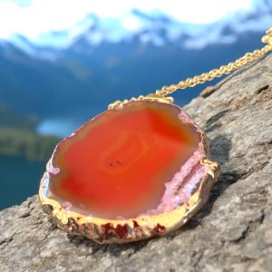 May include: A gold chain necklace with a large, orange agate pendant. The pendant has a natural, irregular shape and a smooth, polished surface.
