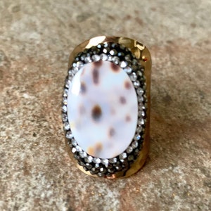 Op de afbeelding: Een gouden ring met een witte ovale schelp cameo ingelegd in een pavé van zwarte strass steentjes.