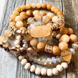 May include: A set of six bracelets featuring various natural materials, including wood, stone, and glass beads. The bracelets are stacked on a wooden surface and include a gold charm with the text "Druzy Rocks".