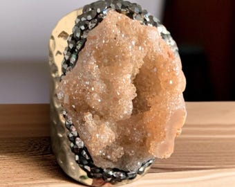 May include: A gold-toned ring with a textured band, featuring a central geode cluster in shades of peach and tan. The geode is surrounded by a band of dark gray crystals, creating a striking contrast. The ring is set on a wooden surface.