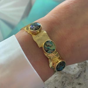 Può includere: Bracciale dorato con tre inserti ovali in conchiglia di abalone iridescente. Il bracciale ha una texture martellata e una forma organica unica. Un gioiello elegante.