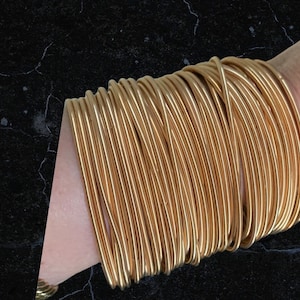 Könnte beinhalten: Ein Stapel goldfarbener Metallarmreifen, die an einem Handgelenk getragen werden.