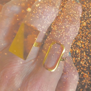 May include: Two gold rings, one rectangular and one oval, worn on a finger. The rings are simple and minimalist in design.