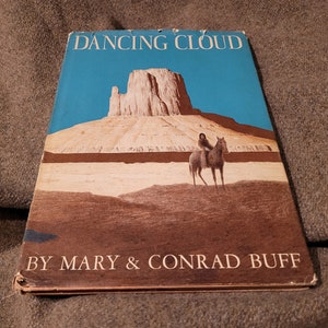 May include: A vintage book titled "Dancing Cloud" by Mary & Conrad Buff. The cover features a colorful illustration of a Native American person riding a horse in front of a large rock formation.