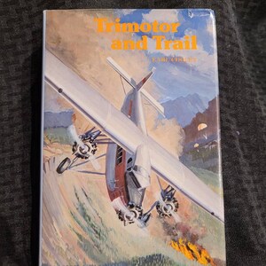 May include: A vintage book cover featuring a white and red airplane flying over a mountain range with a fire in the foreground. The book title is "Trimotor and Trail" by Earl Coopey.