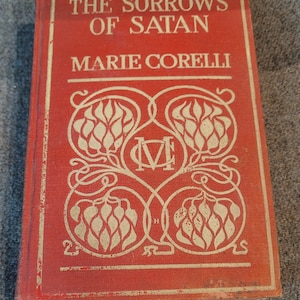 May include: A vintage book titled "The Sorrows of Satan" by Marie Corelli. The cover is a faded red with white ornate floral designs and text. The book's title and author's name are prominently displayed.