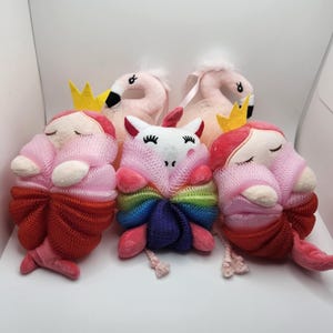 May include: A collection of plush bath accessories, including two flamingo-shaped items with yellow crowns, and three doll-like figures with closed eyes. The dolls have pink and red mesh bodies and bows.