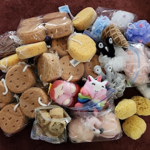 May include: A collection of bath accessories including round yellow sponges, mesh bath poufs in various colors, and plush character-shaped sponges including a pink unicorn, a red-haired figure, and ice cream cone designs, all individually packaged.