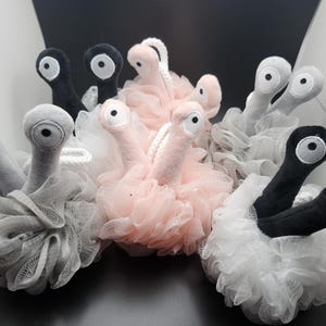 May include: A collection of whimsical bath sponges in various colors, including pink, gray, black, and white. Each sponge features a plush, eye-shaped design on top of a mesh loofah base, creating a playful aesthetic.