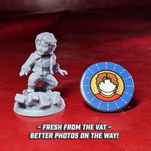 May include: A gray 3D printed miniature figure of a person with brown hair and a red shirt. The figure is standing on a gray rock base. A blue and yellow button with a cartoon image of a person with brown hair and a red shirt is also shown.
