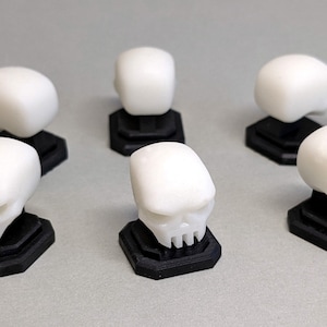 May include: Six white skull figurines on black bases. The skulls have a detailed design and are made of a smooth, white material.
