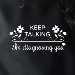 May include: Black sweatshirt with white text that reads "Keep Talking I'm Diagnosing You" with floral accents.