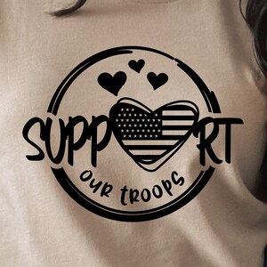 May include: A tan t-shirt with a black circle design featuring a heart with an American flag pattern and the text "Support Our Troops".