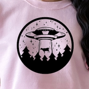 Könnte beinhalten: Ein rosa Sweatshirt mit einer schwarzen Grafik eines UFOs, das eine Kuh in einem Wald entführt. Die Grafik ist von Sternen umgeben.