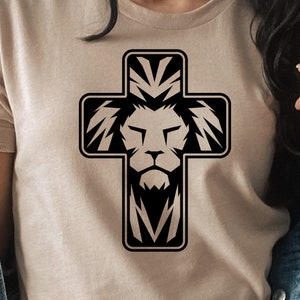 May include: A beige t-shirt featuring a black graphic of a cross with a lion's face at the center. The lion's mane and the cross's arms are stylized with sharp, angular lines. The shirt has short sleeves and a crew neck.