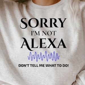 以下が含まれることがあります： 「SORRY I'M NOT ALEXA」の黒い文字、青い波形グラフィック、黒い文字で「DON'T TELL ME WHAT TO DO！」がプリントされたライトグレーのスウェットシャツ。スウェットシャツは柔らかい素材で作られています。