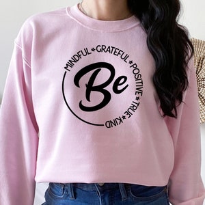 May include: Pink sweatshirt with a black circle graphic that says "Be" with the words "Mindful", "Grateful", "Positive", "True", and "Kind" around the circle.