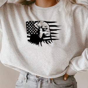 Pu&ograve; includere: Una felpa grigio chiaro con una grafica in bianco e nero di una bandiera americana e la testa di un'aquila. La testa dell'aquila &egrave; sul lato destro del disegno della bandiera.