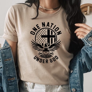 Pu&ograve; includere: Una maglietta beige con un design grafico nero che presenta un'aquila con le ali spiegate, una croce e le parole "One Nation Under God" in un cerchio.