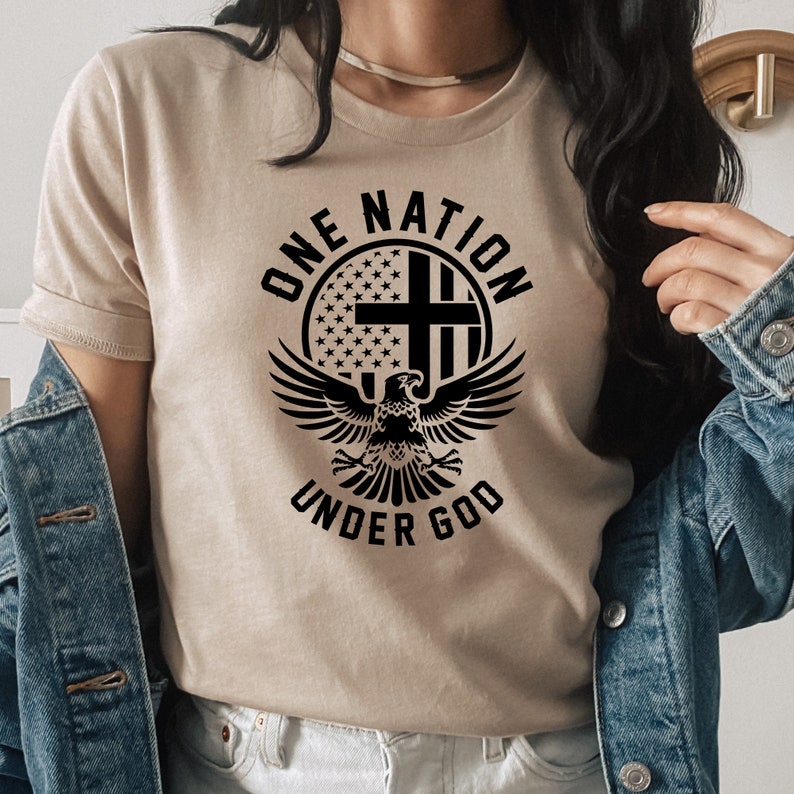 Pu&ograve; includere: Una maglietta beige con un design grafico nero che presenta un'aquila con le ali spiegate, una croce e le parole "One Nation Under God" in un cerchio.