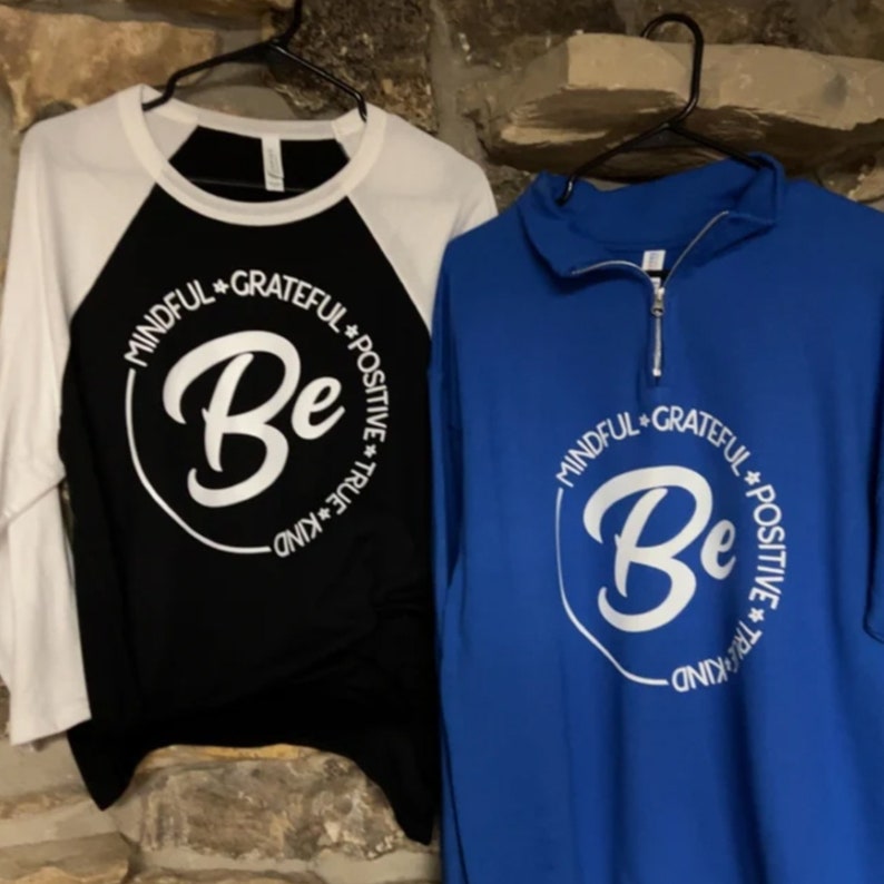 May include: Two long-sleeved shirts with a white circle design that says "Be" with the words "Mindful", "Grateful", "Positive", "True", and "Kind" around the circle. The shirt on the left is black with white sleeves and the shirt on the right is blue.