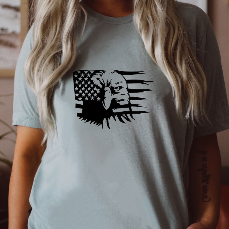 Pu&ograve; includere: T-shirt azzurra con grafica in bianco e nero di una testa d'aquila e della bandiera americana. La testa dell'aquila &egrave; a destra, con le stelle e le strisce della bandiera a sinistra.