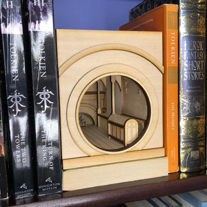 May include: A wooden bookend shaped like a doorway from The Lord of the Rings. The bookend features a cutout of a hobbit hole interior. The bookend is displayed with a collection of The Lord of the Rings books.