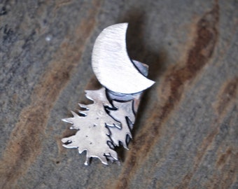 Puede incluir: Un colgante de plata con una luna creciente y un diseño de árbol estilizado. El colgante está hecho de metal y tiene una superficie texturizada.