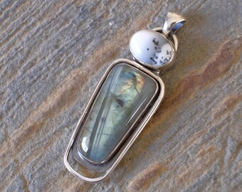 Puede incluir: Un colgante de plata con una gran piedra de labradorita rectangular e iridiscente engastada en un marco de plata. Una piedra de ágata más pequeña, redonda, blanca y gris está engastada sobre la labradorita.