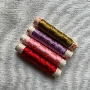 May include: Four spools of embroidery floss in red, pink, purple, and gold. The spools are wrapped in white paper with red text.