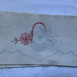 Könnte beinhalten: Cremefarbener Stoff mit einem gestickten Blumenmuster. Eine rote Gänseblümchen und ein Korb mit Blumen in blauem Faden sind abgebildet. Der untere Rand hat eine blau geschwungene Bordüre. Der Stoff ist gefaltet.