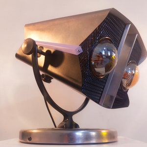 Puede incluir: Una lámpara de mesa de metal negro y plateado con dos bombillas expuestas y una base de metal negro. La lámpara tiene un diseño único con una forma geométrica y un patrón de rejilla en la parte delantera.